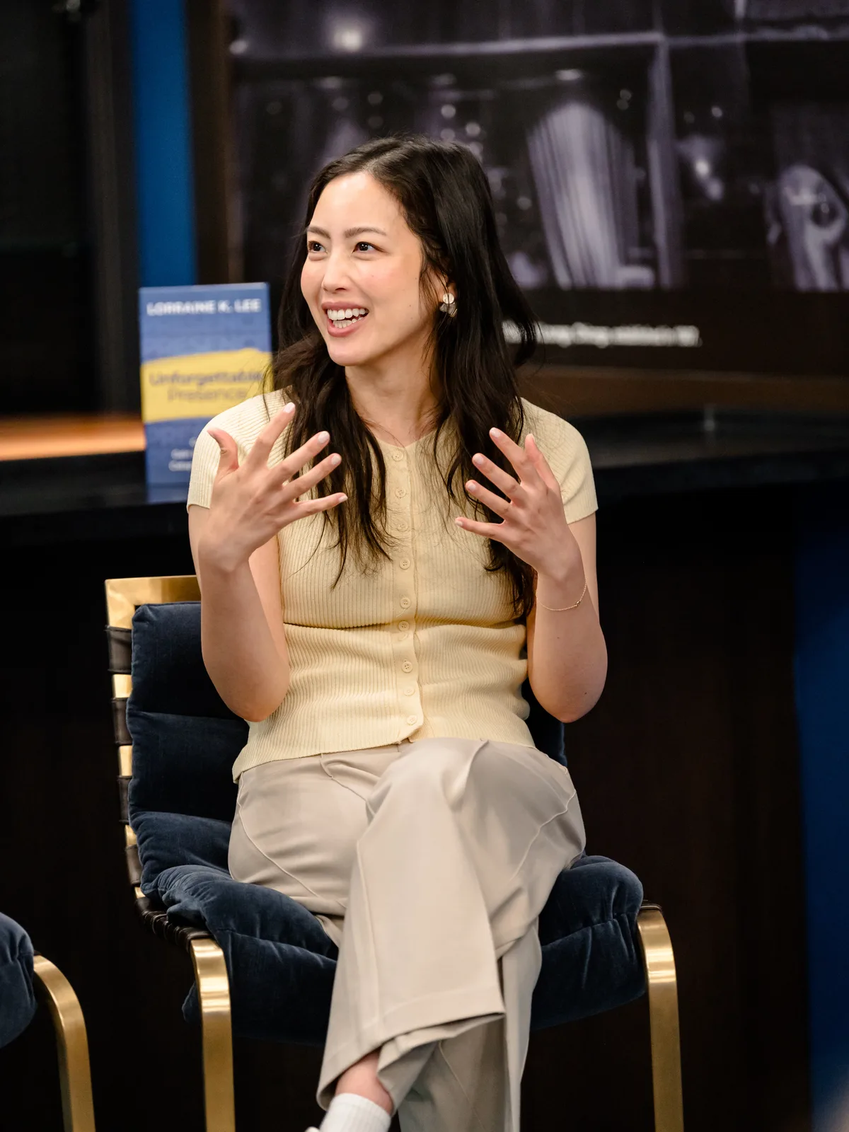 Lorraine K. Lee seated in a studio interview, with her book Unforgettable Presence displayed beside her.